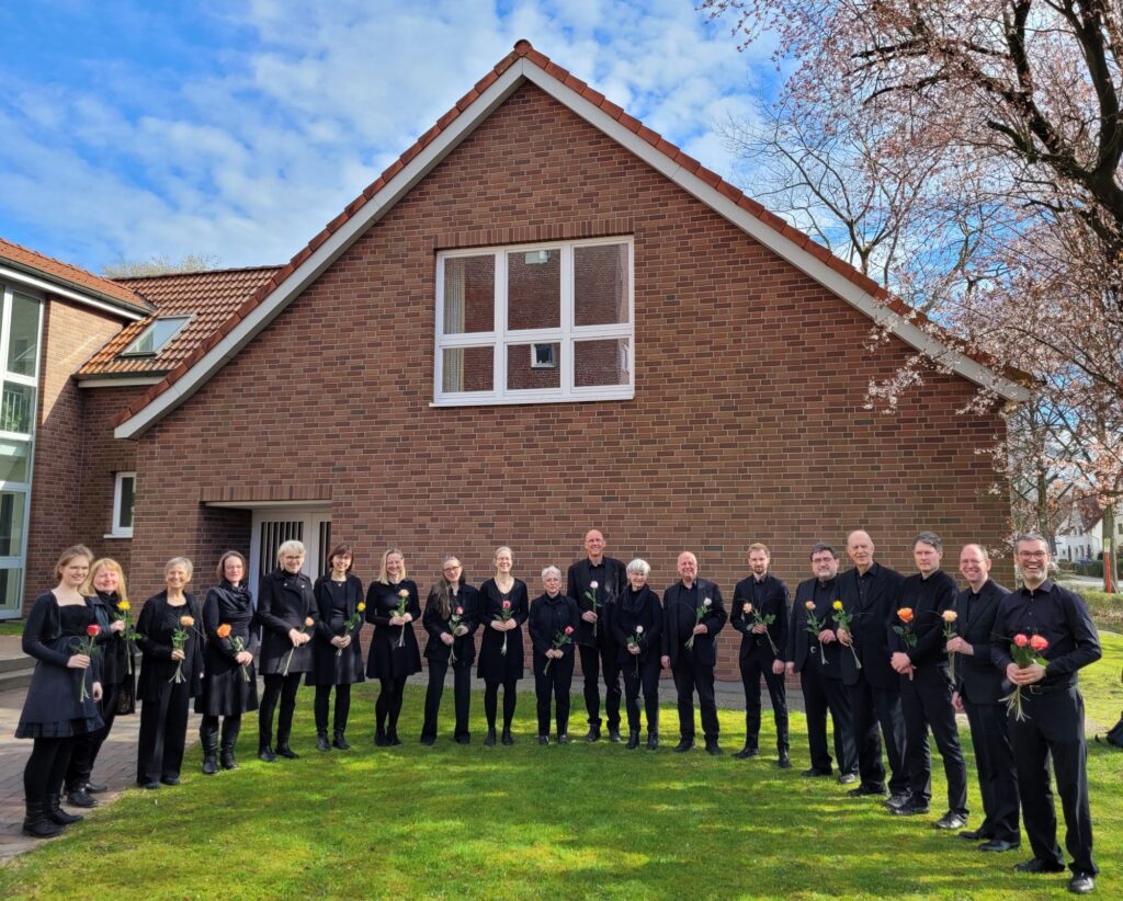 Unser Chor in Hamburg-Wandsbek steht in schwarzer Chorkleidung mit Rosen in der Hand im Freien. Rechts ist ein blühender Baum zu sehen.