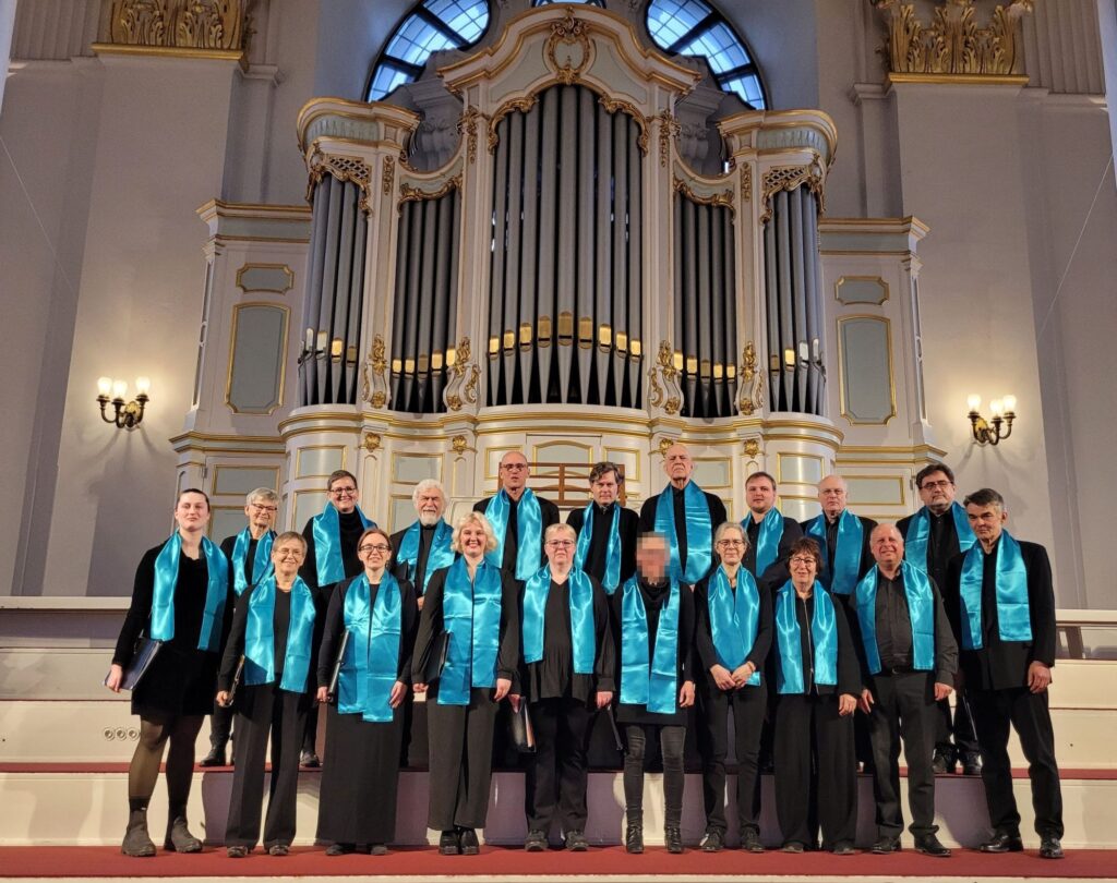 Der Wandsbeker Kammerchor steht in schwarzer Chorkleidung mit türkisfarbenen Schals vor einer Orgel.